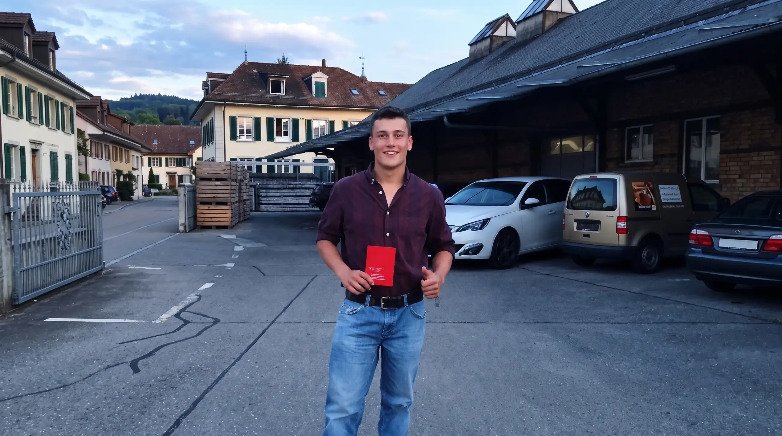 Junger Mann in Jeans und Hemd hält Buch in Hofeinfahrt mit parkenden Autos und historischen Gebäuden im Hintergrund.
