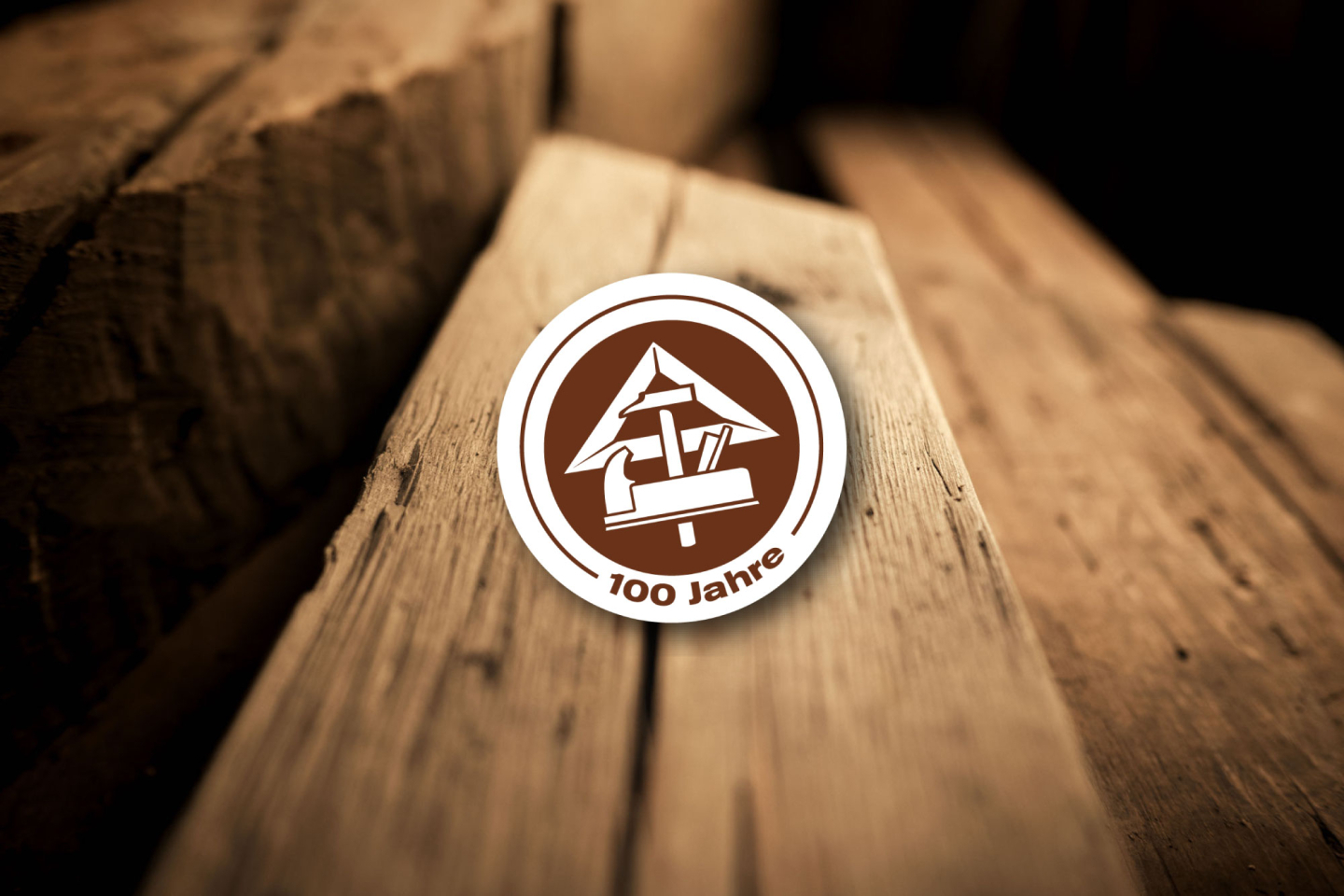 Holzstapel im Hintergrund, rundes Logo mit Werkzeugen und 100 Jahre Schriftzug im Vordergrund.